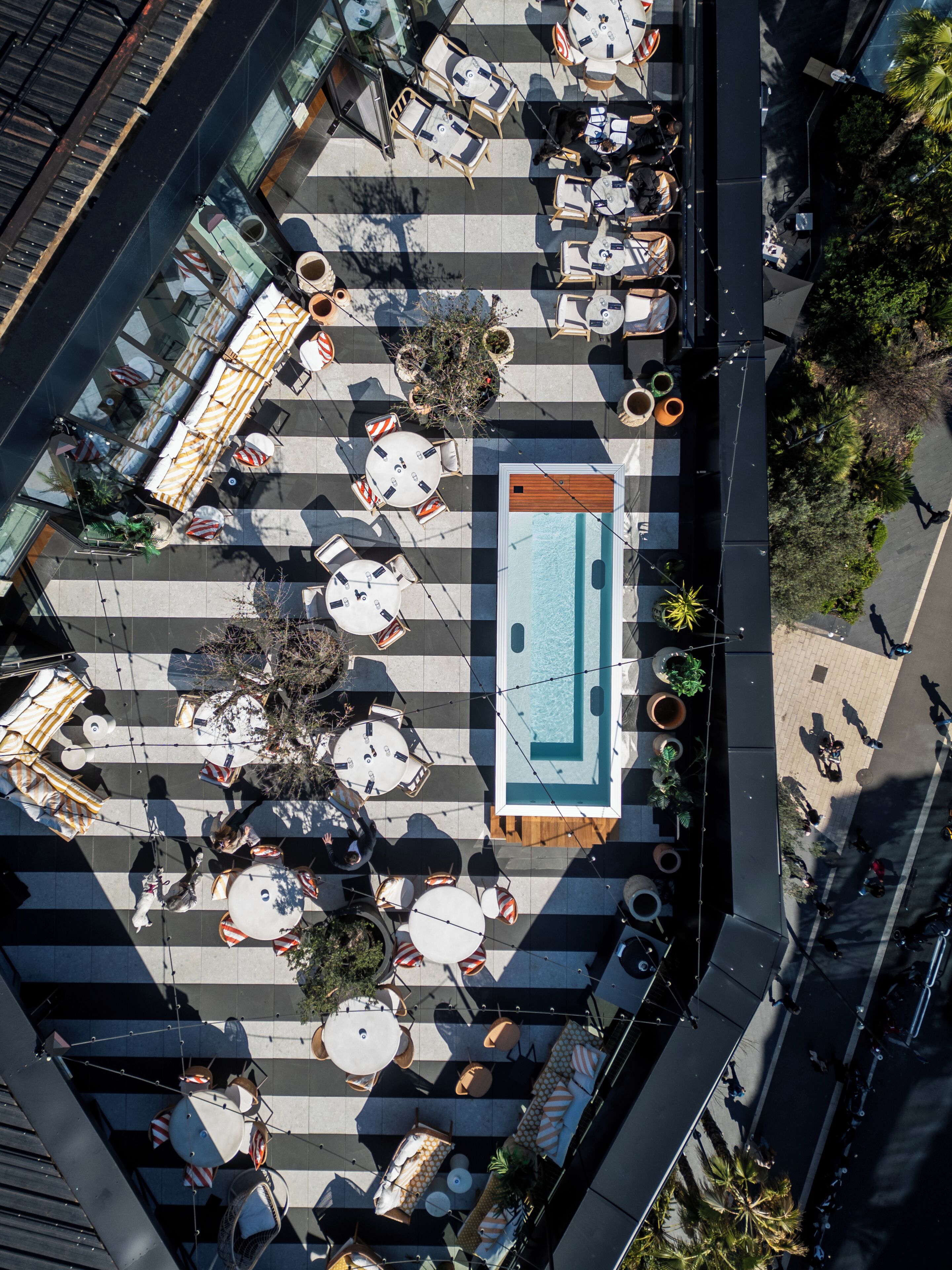 An aerial view of a rooftop terrace with multiple tables and chairs, a swimming pool, and surrounding greenery.