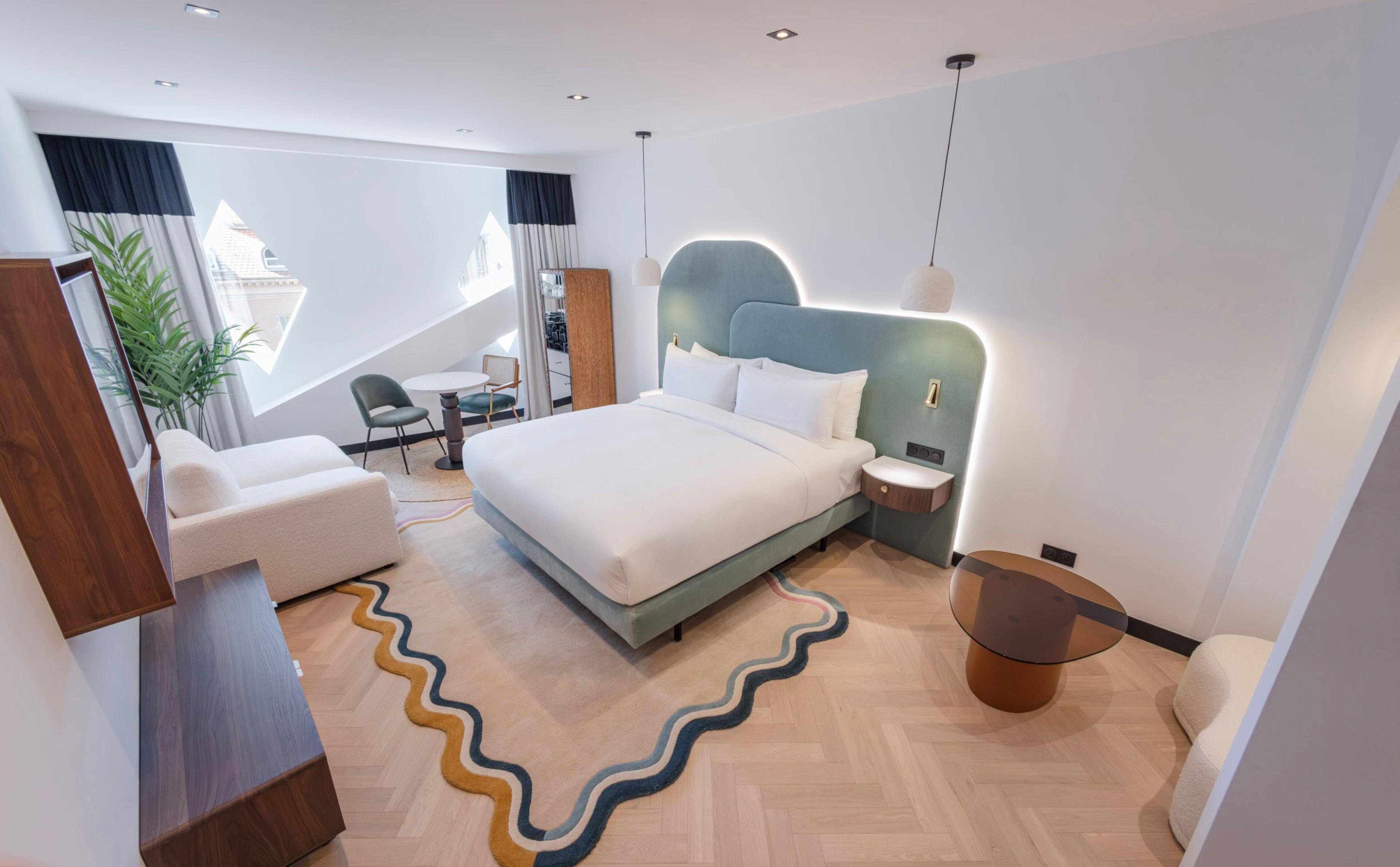 A modern hotel room with a bed, bedside table, lamp, and a view of the city.
