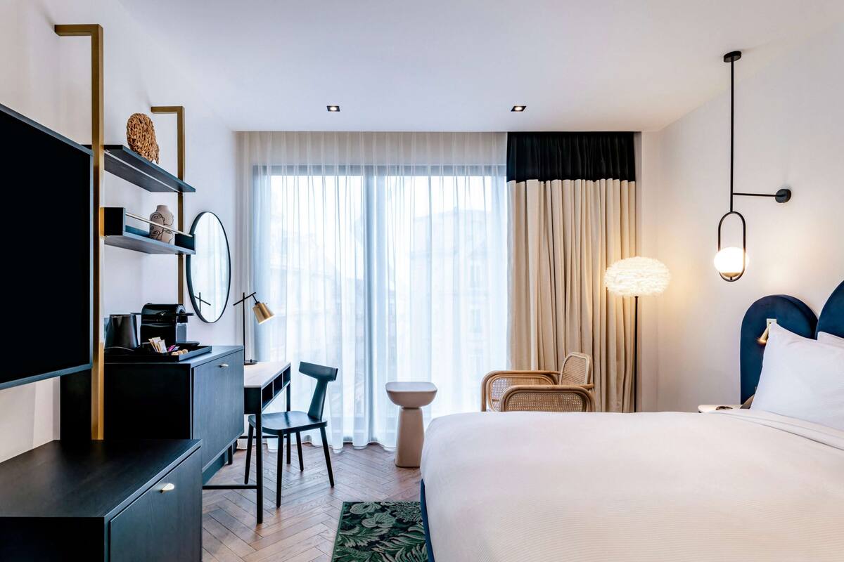 A hotel room with a bed, a desk, a chair, a TV, a lamp, and a window with curtains.