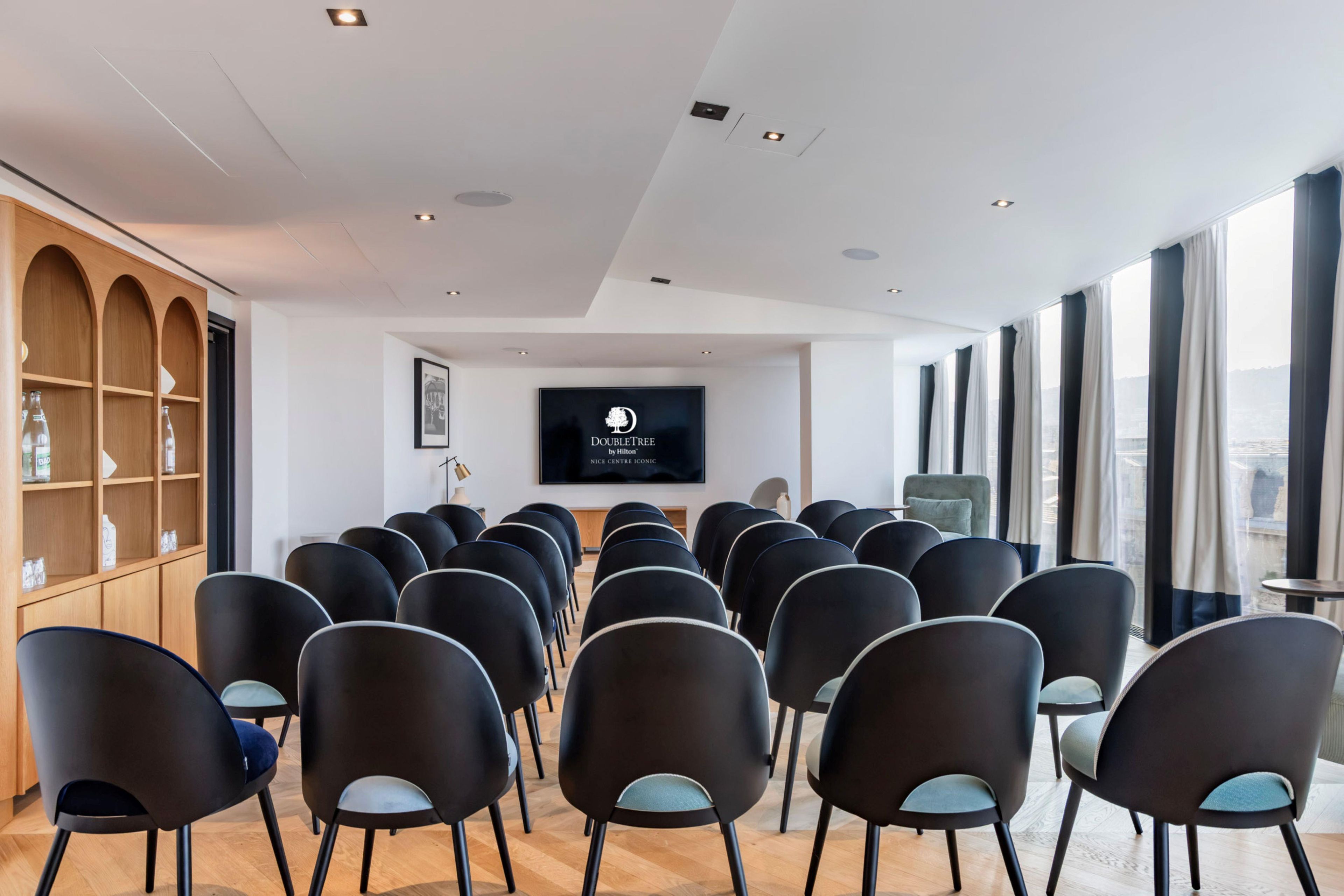 Un salón de conferencias con filas de sillas mirando hacia una pantalla, estanterías de madera y grandes ventanales con cortinas.