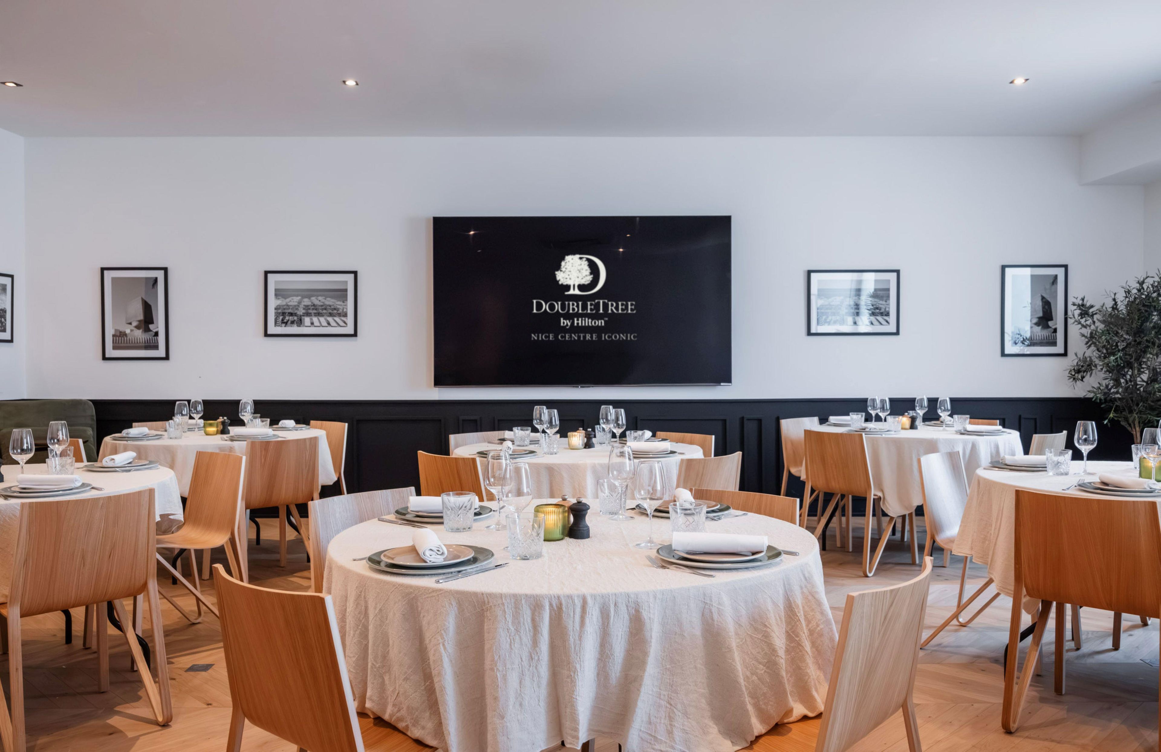 Een eetgedeelte met ronde tafels gedekt voor een maaltijd, een groot scherm waarop het logo van DoubleTree by Hilton staat, en kunstwerken in lijsten aan de muur.