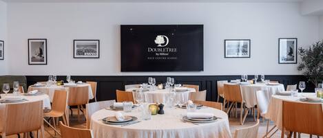 A dining area with round tables set for a meal, a large screen displaying the DoubleTree by Hilton logo, and framed artwork on the walls.