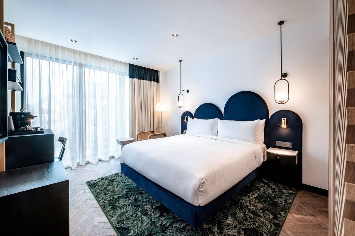 A modern hotel room with a large bed, a desk, and a chair. There are two bedside tables with lamps, a large window with curtains, and a view of the city.