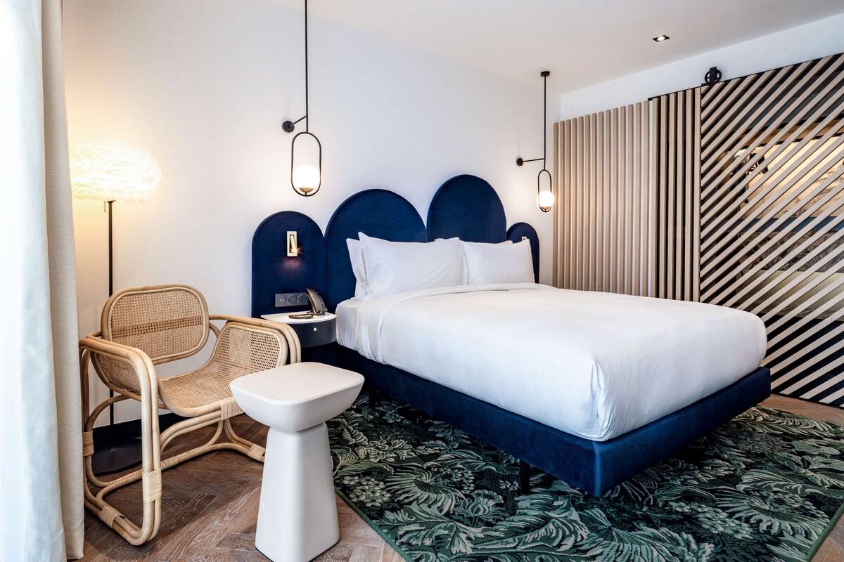 A modern hotel room with a large bed, a bedside table, a chair, and a floor lamp.