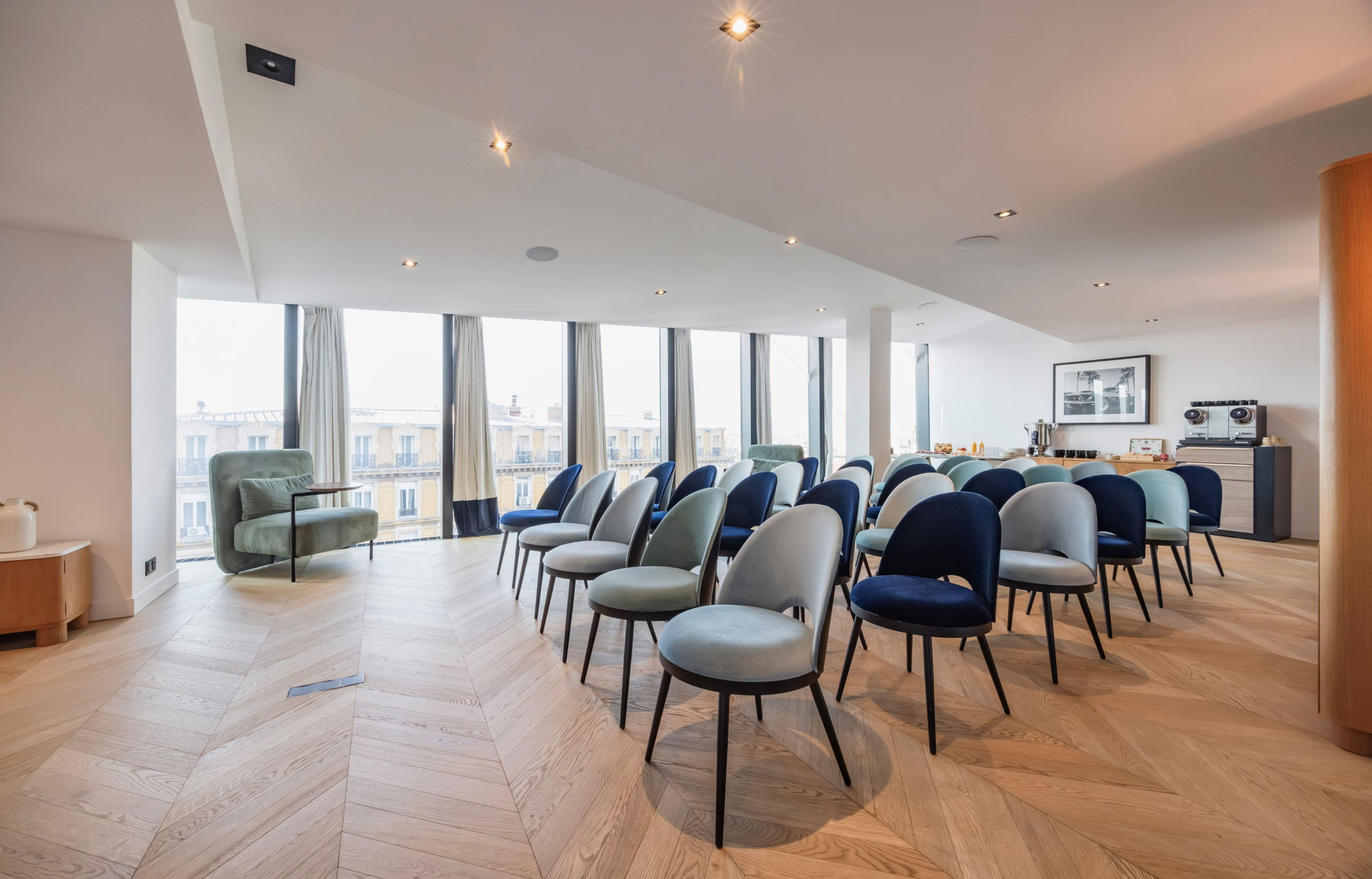A spacious room with a modern design, featuring a large seating area with multiple chairs, a wooden herringbone floor, and large windows offering a view of the cityscape.