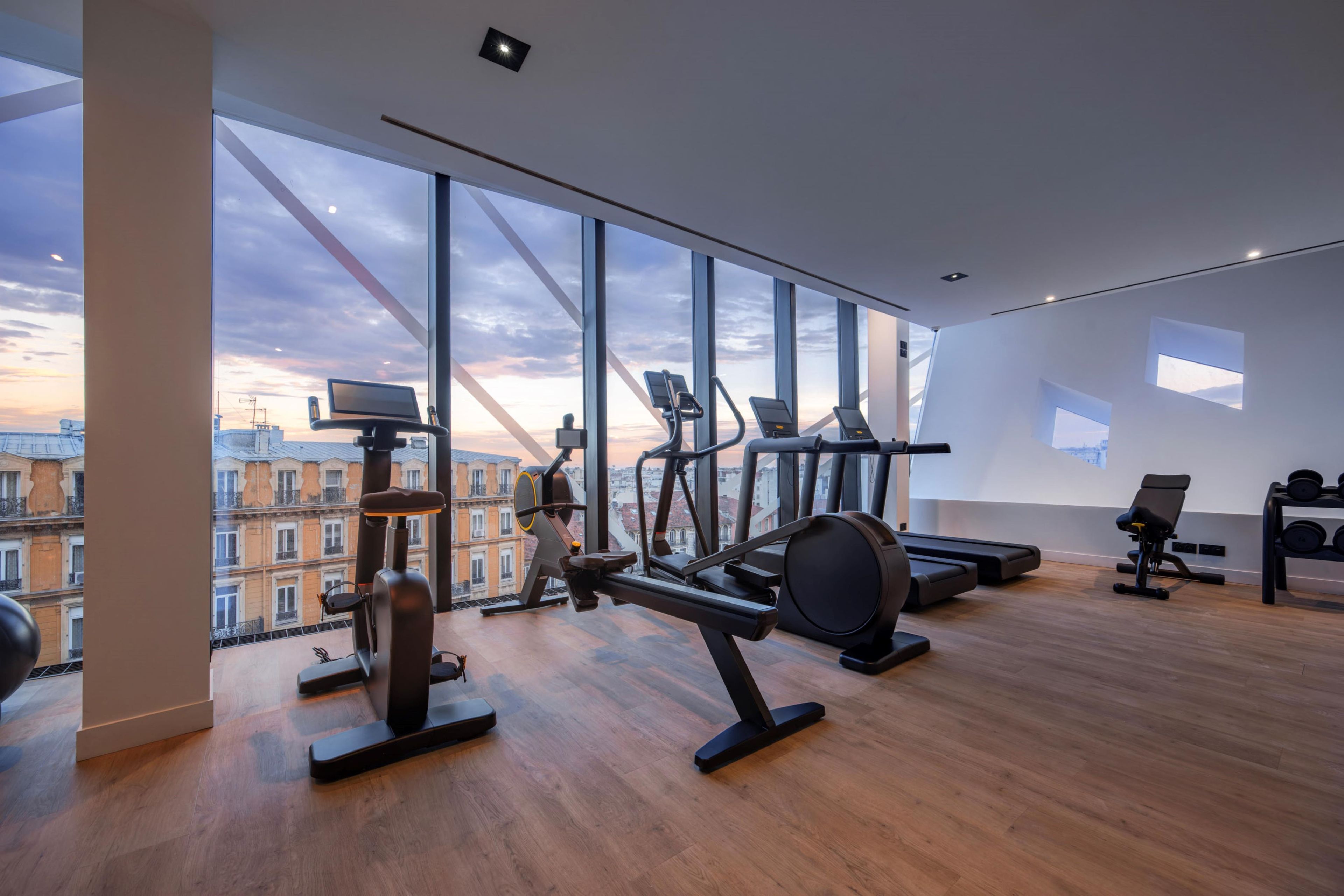 A modern gym with large windows, exercise equipment, and a view of buildings.