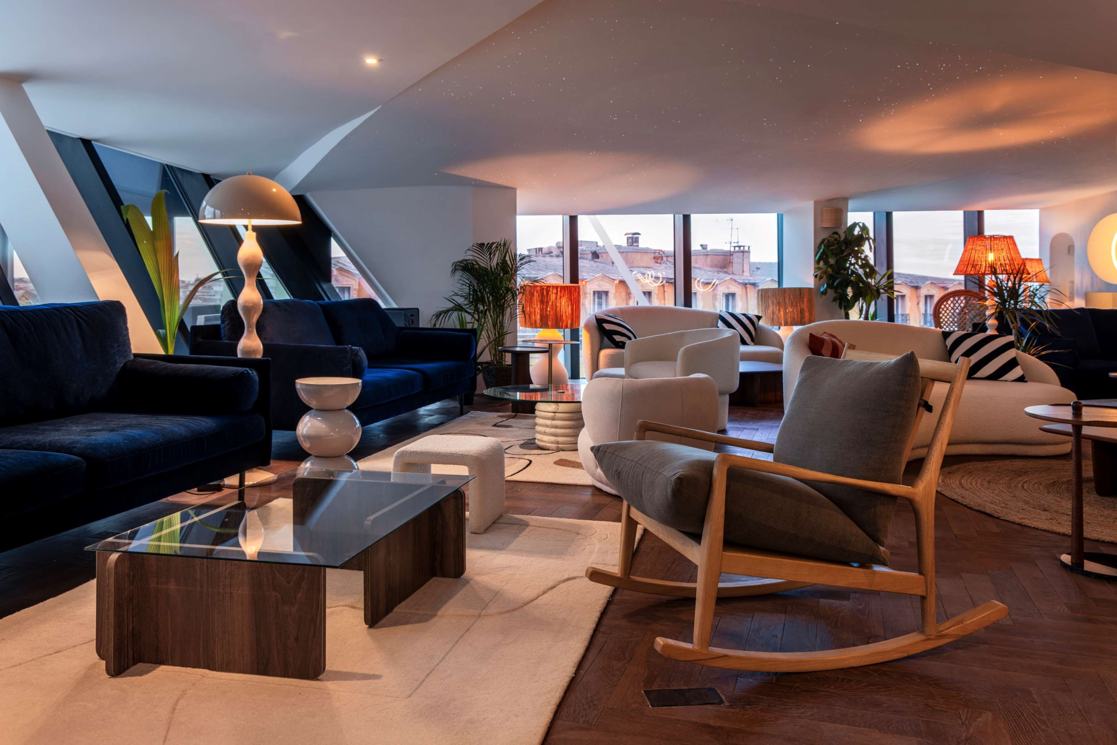 Un salon moderne avec un canapé bleu foncé, une table basse en verre, un fauteuil à bascule en bois et une vue sur la ville grâce à de grandes fenêtres.