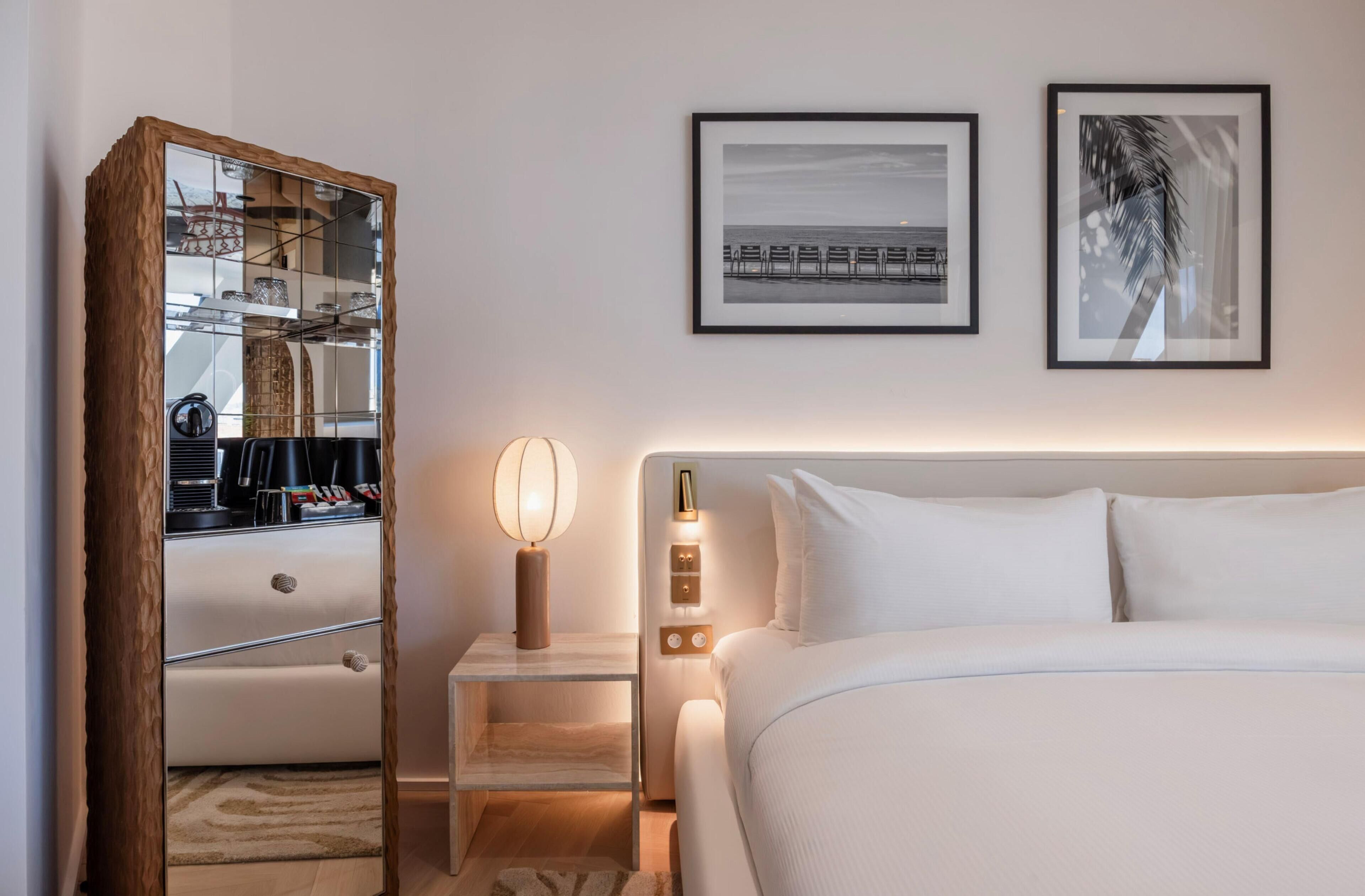 Una habitación de hotel con cama, mesita de noche, lámpara y armario con espejo. Hay dos cuadros en la pared.