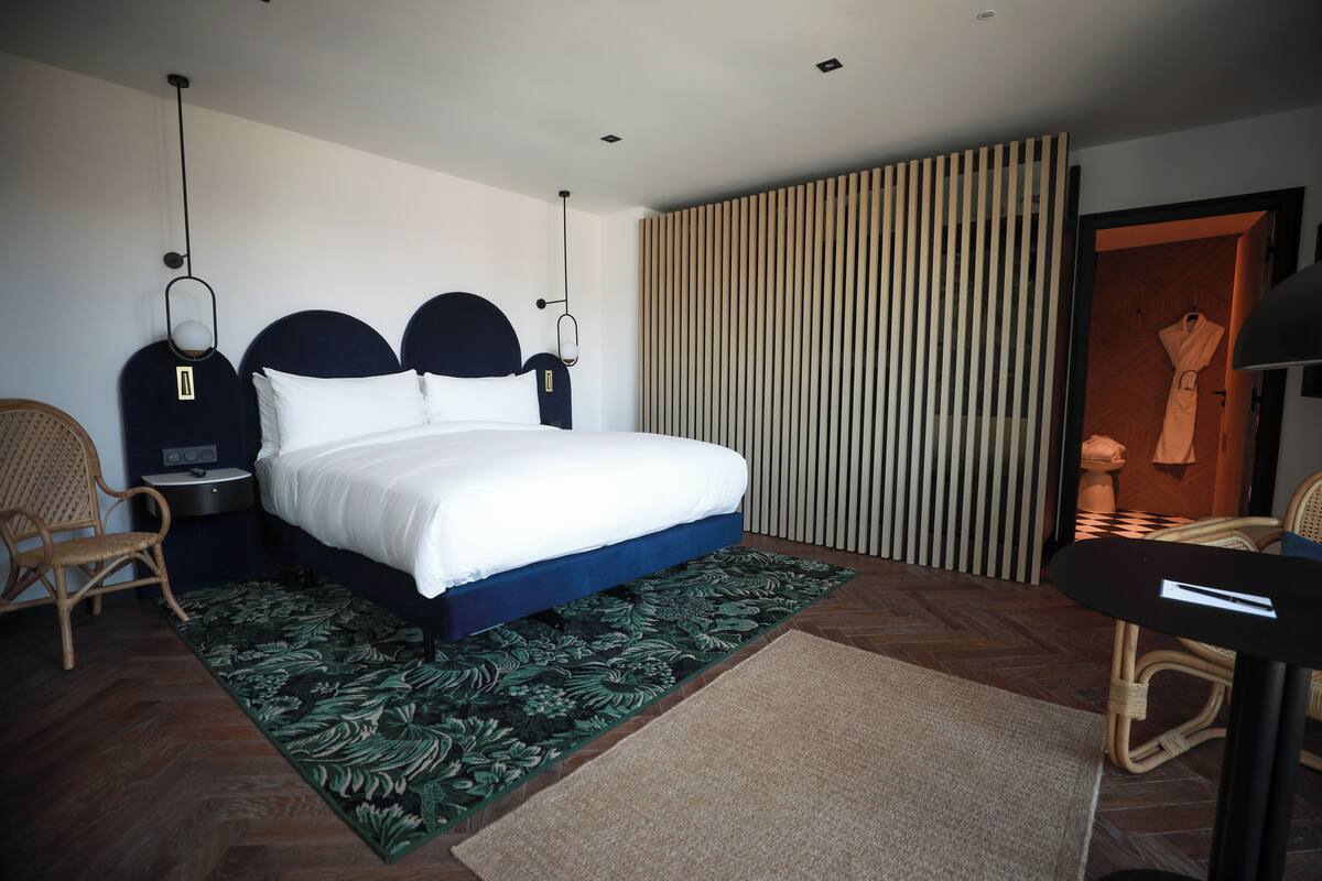 A modern hotel room with a large bed, a side table, a chair, a wardrobe, and a large window with blinds.