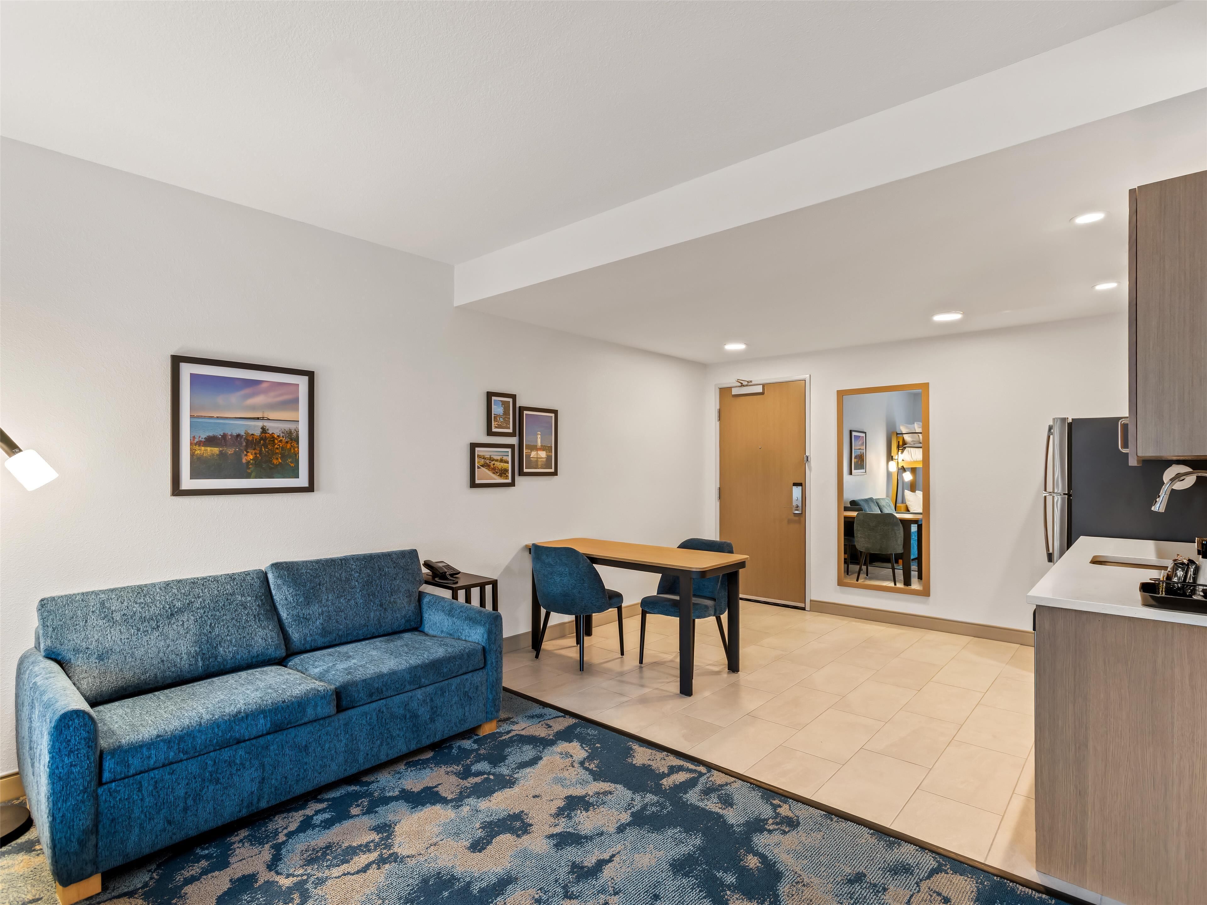 Una habitación de hotel moderna con un sofá azul, una mesa de comedor con sillas, una cocina con refrigerador y un espejo grande.