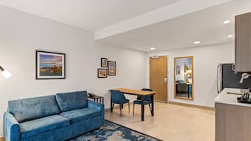 Una habitación de hotel moderna con un sofá azul, una mesa de comedor con sillas, una cocina con refrigerador y un espejo grande.