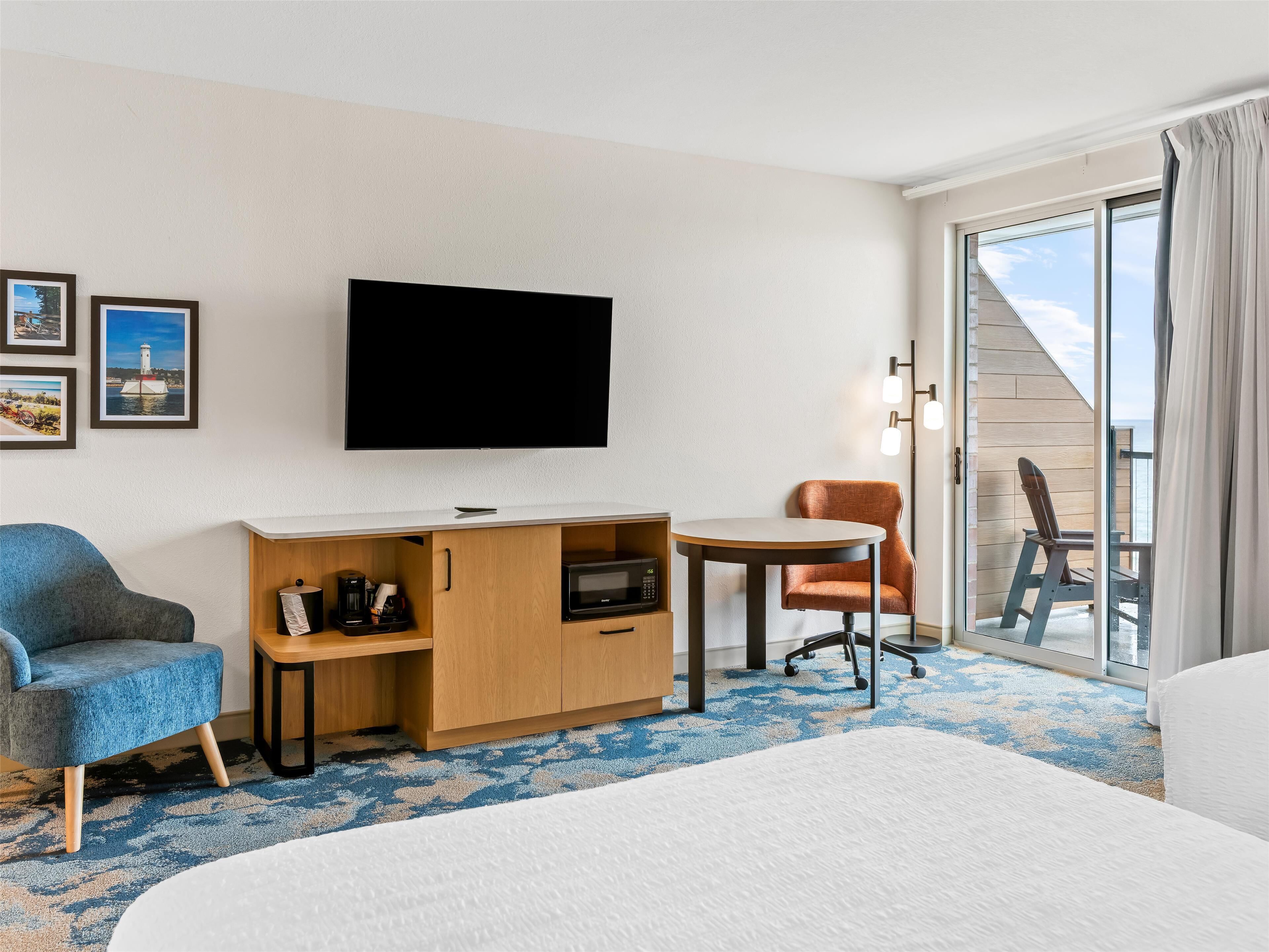A hotel room with a bed, a desk with a flat-screen TV, a blue armchair, an orange chair, and a view of the sea through a sliding glass door.