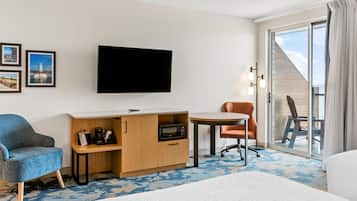 A hotel room with a bed, a desk with a flat-screen TV, a blue armchair, an orange chair, and a view of the sea through a sliding glass door.