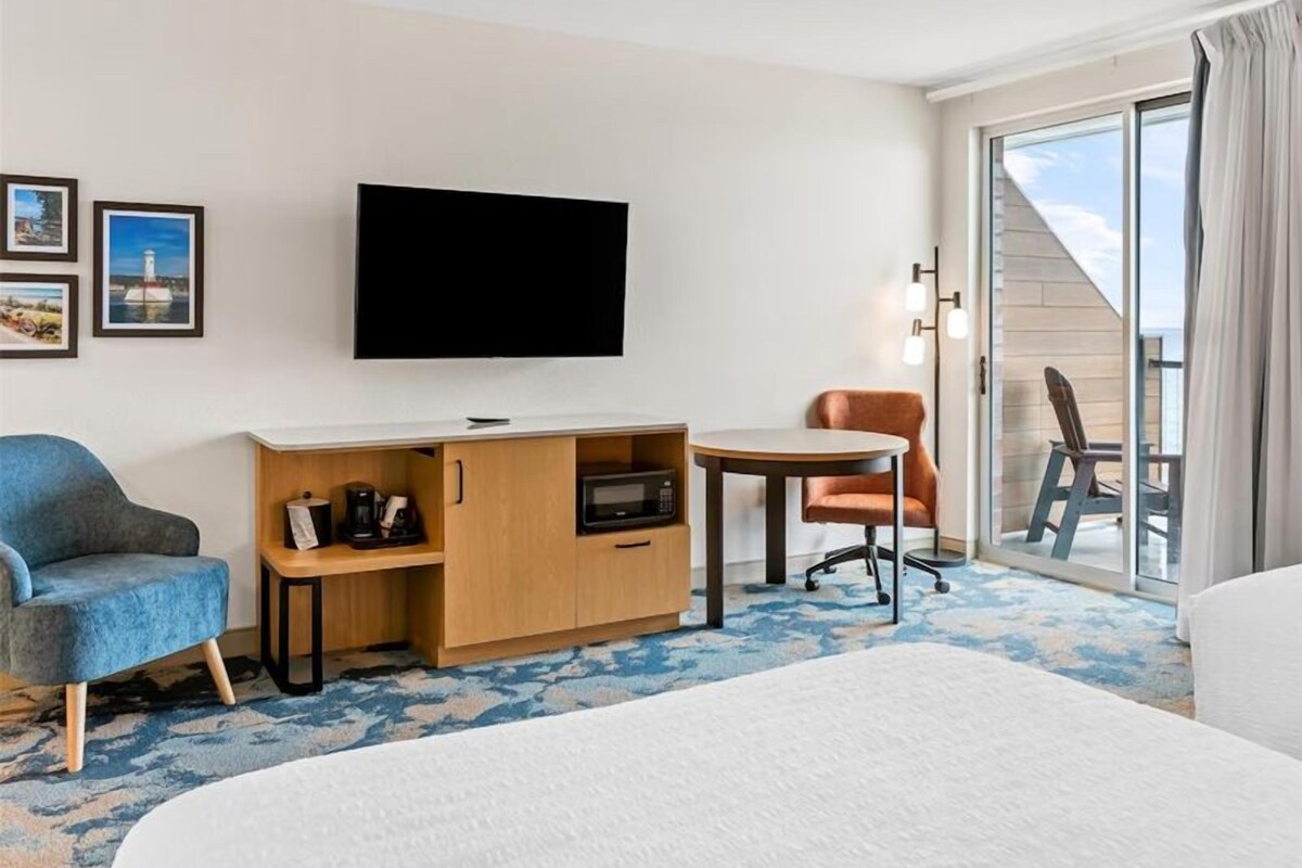 A hotel room with a bed, a desk with a flat-screen TV, a chair, a small table, and a view of the outside.