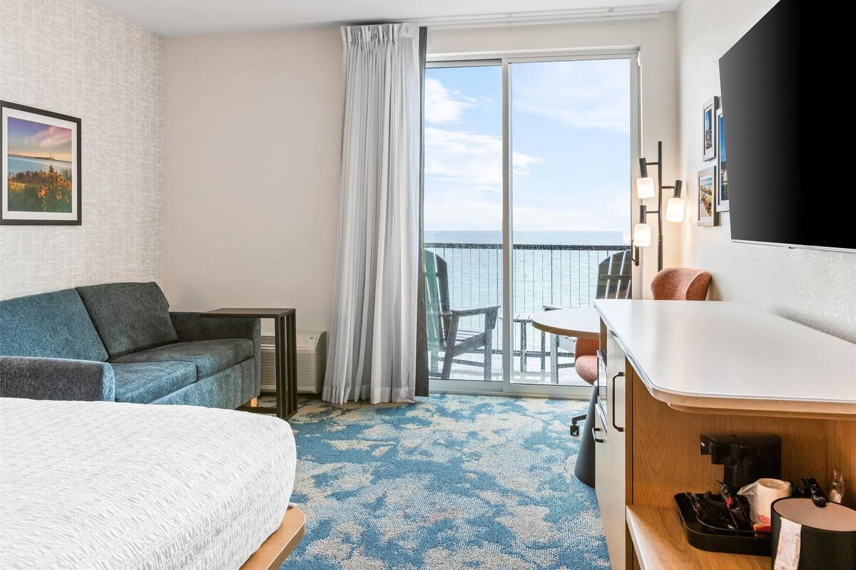 A hotel room with a bed, a sofa, a desk, a chair, a coffee machine, and a balcony with a view of the sea.