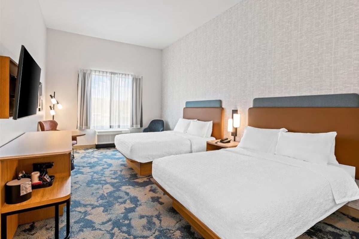 Quarto de hotel com duas camas, uma escrivaninha, uma cadeira, televisão e janela com cortinas.