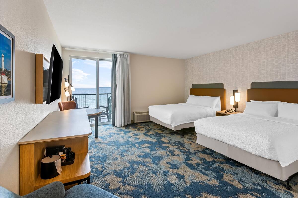 A hotel room with two beds, a desk, a chair, a TV, and a view of the sea.
