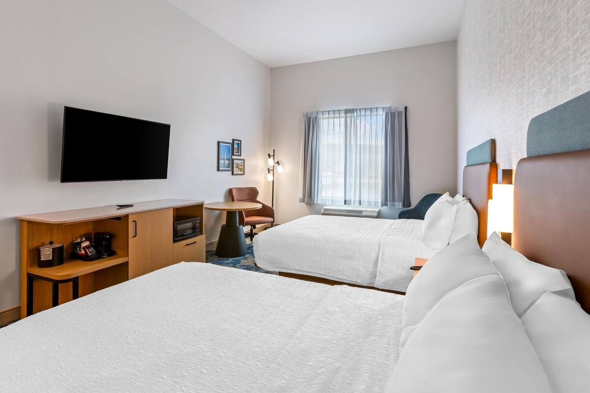 A hotel room with two beds, a desk with a microwave, a chair, a TV, and a window with curtains.