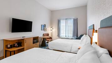 Ein Hotelzimmer mit zwei Betten, einem Schreibtisch mit Mikrowelle, einem Sessel, einem Fernseher und einem Fenster mit Vorhängen.