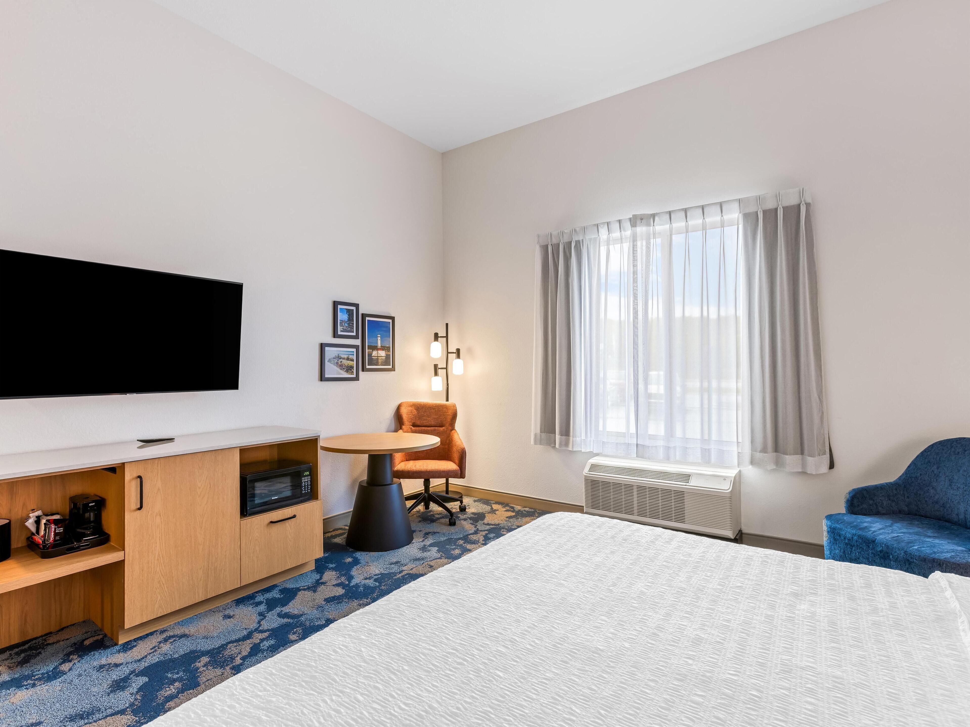 A hotel room with a flat-screen TV, a wooden cabinet with a microwave, a round table with an orange chair, a blue sofa, and a large window with sheer curtains.
