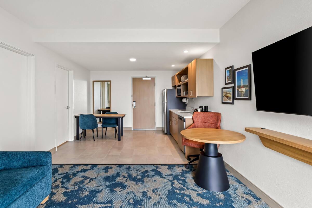 A modern hotel room with a dining area, kitchenette, and a large TV.