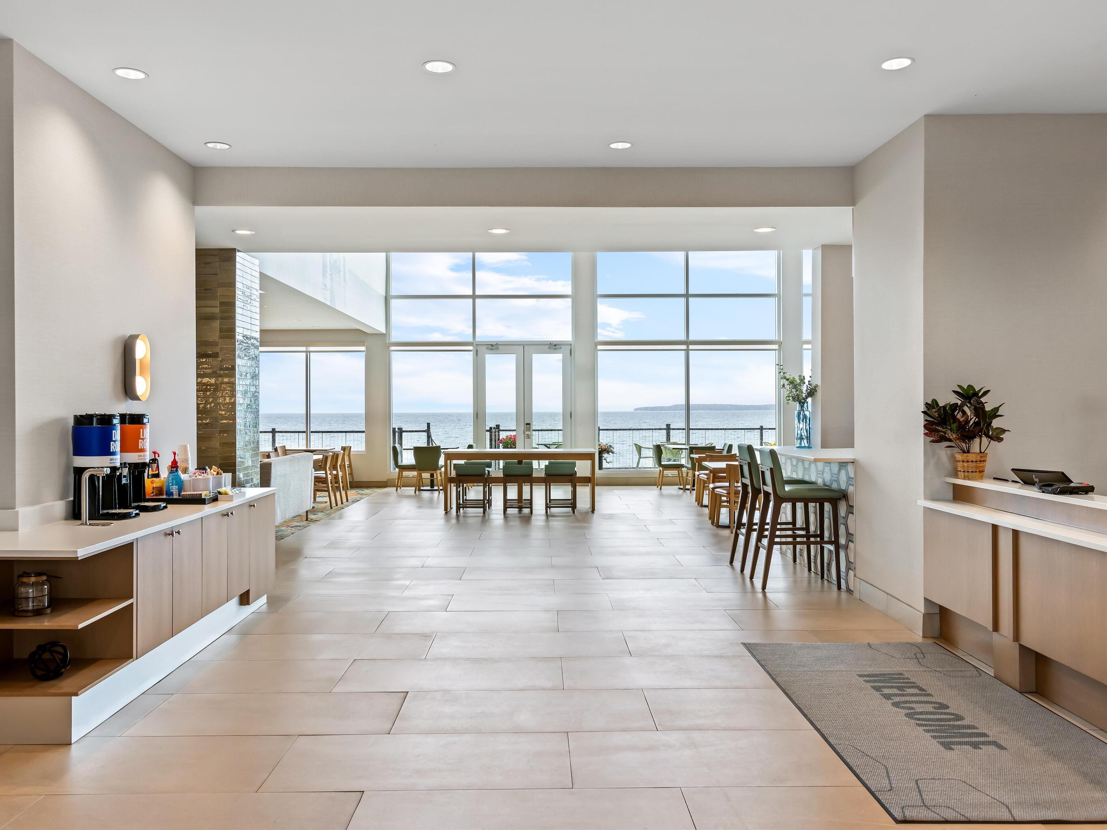 A spacious hotel lobby with a large seating area, a coffee station, and a view of the sea through large windows.