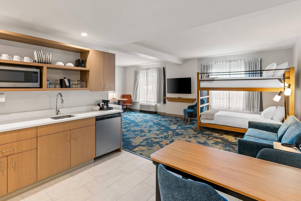 A modern hotel room with a kitchenette, bunk beds, a flat-screen TV, and a cozy seating area.
