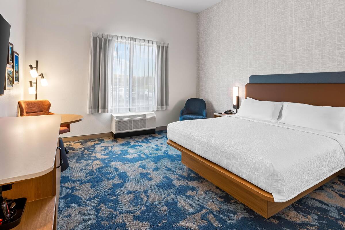 A hotel room with a large bed, a desk, a chair, a window with curtains, a wall-mounted air conditioner, and a patterned carpet.