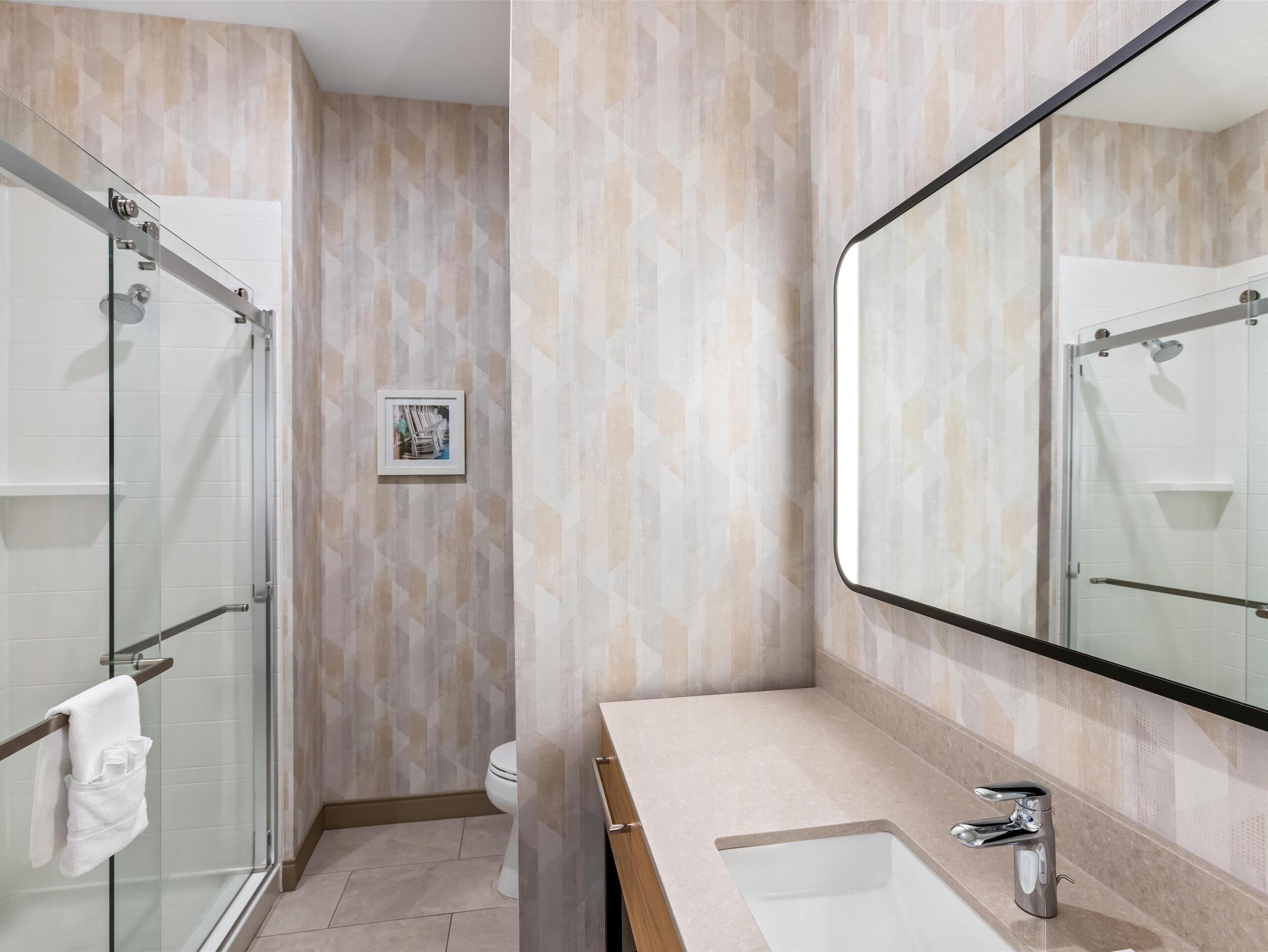 Un baño moderno con cabina de ducha de vidrio, un espejo grande y pared con azulejos color beige.