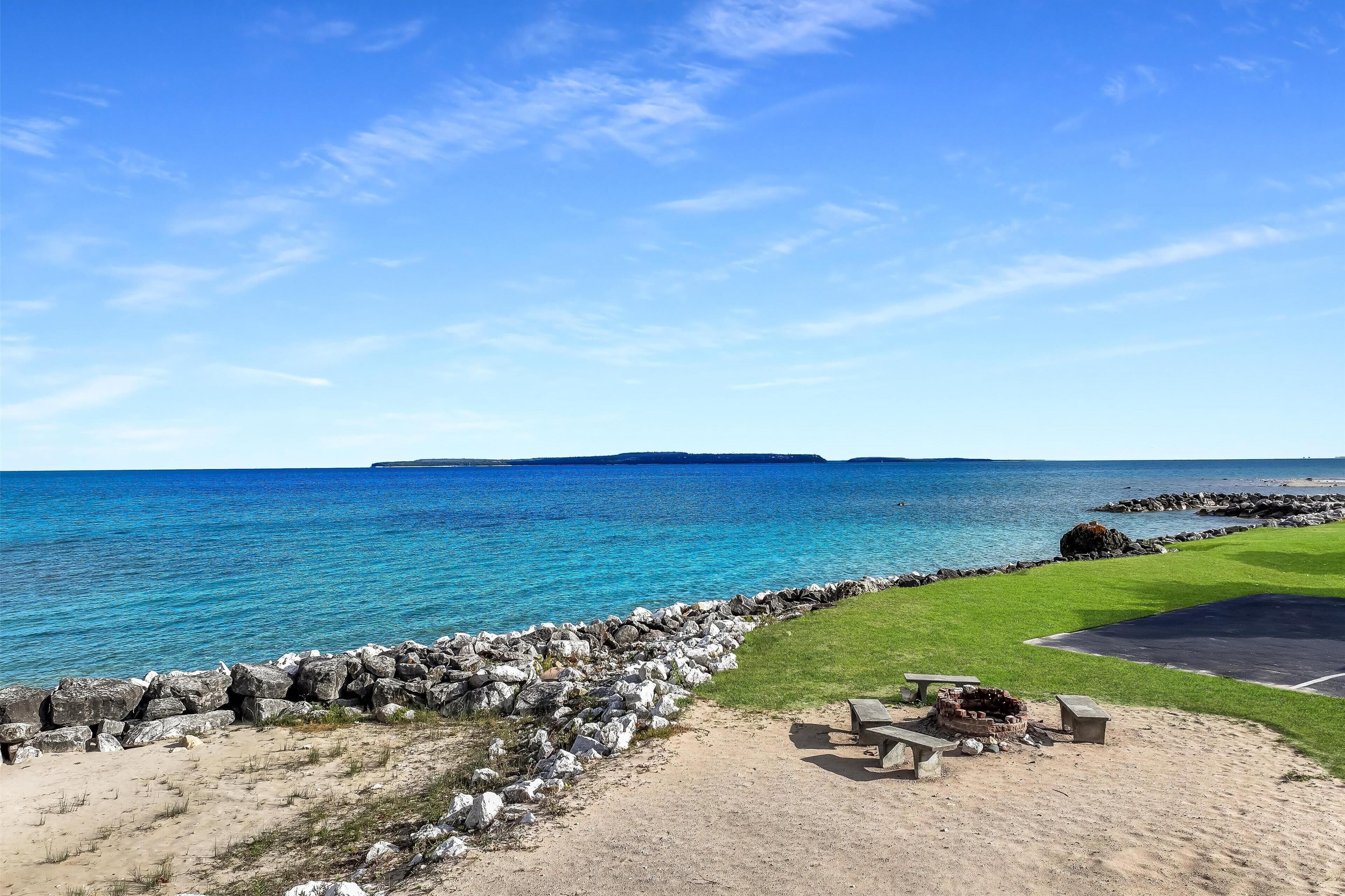 Une plage avec une digue rocheuse, une zone herbeuse dotée d’un foyer, et une vue sur la mer et la terre au loin.