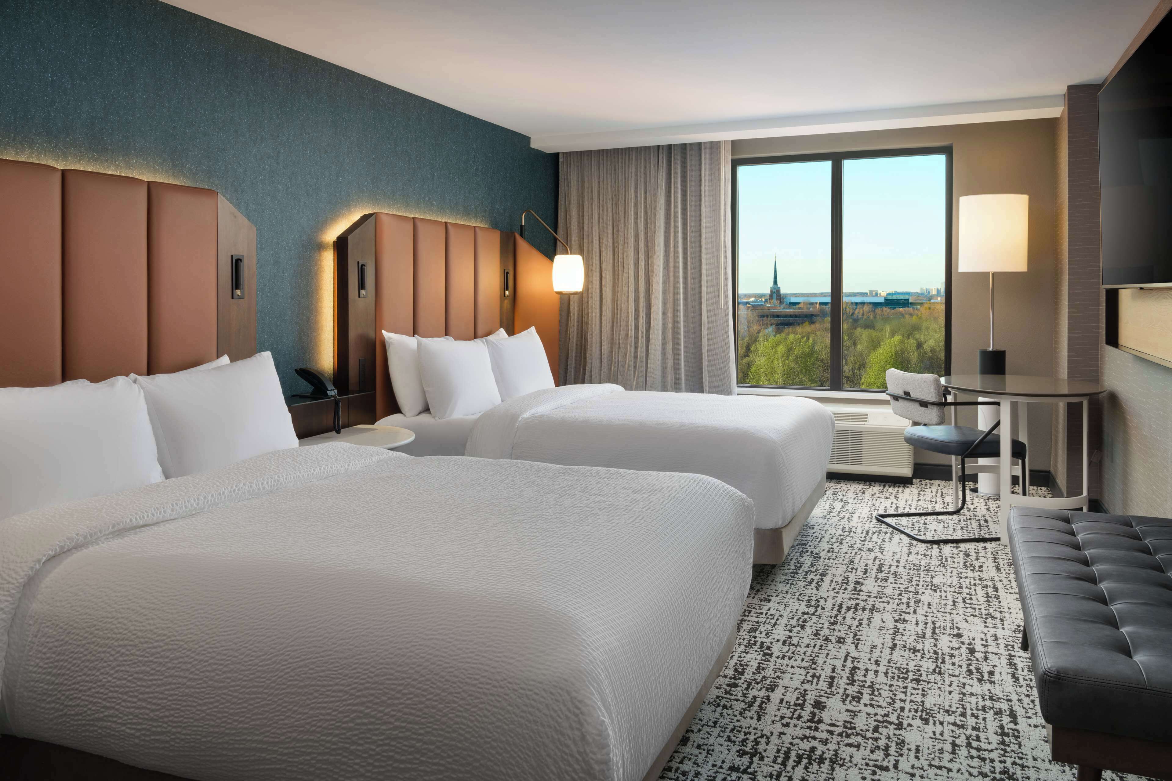 A hotel room with two beds, a desk, a chair, a sofa, and a large window offering a city view.