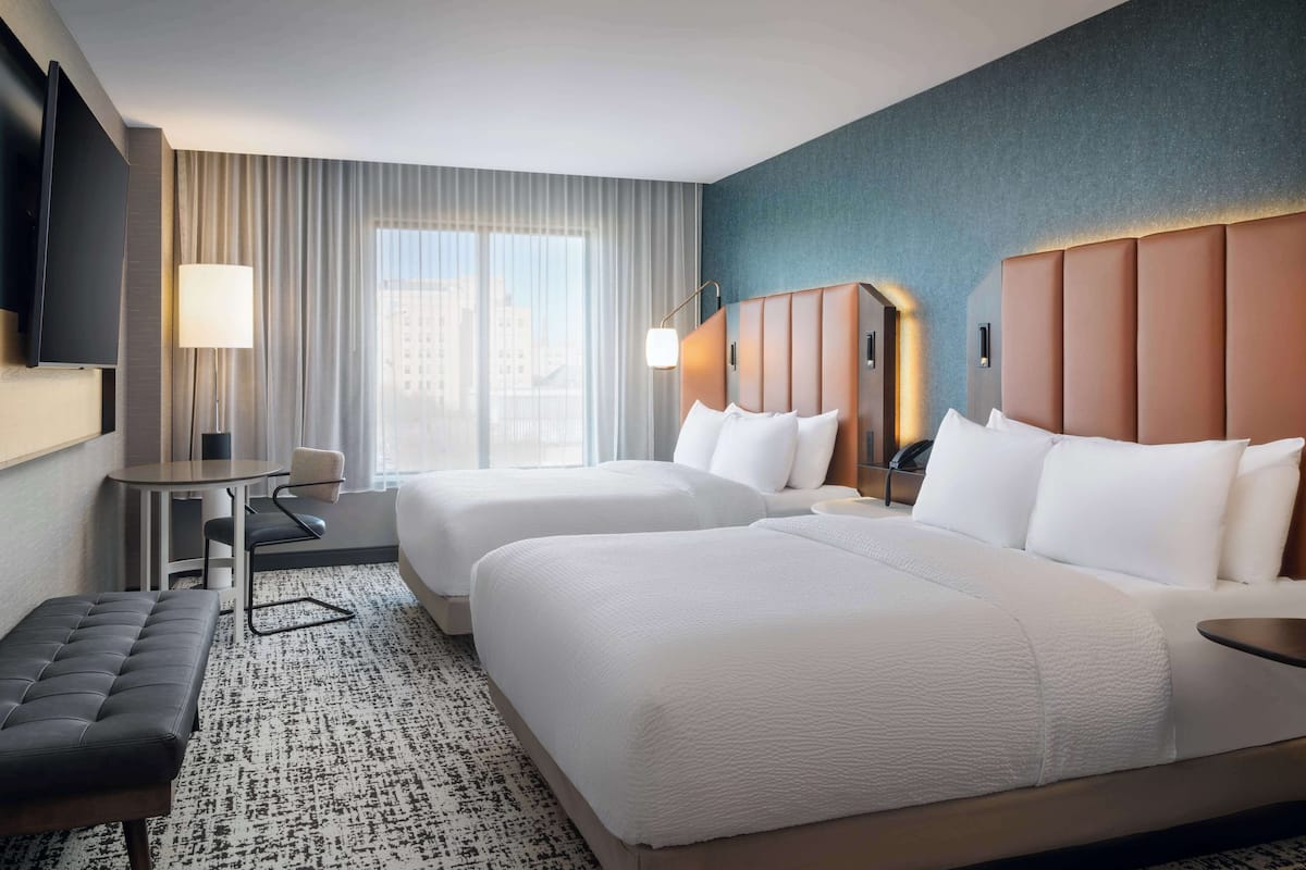 A hotel room with two beds, a desk, a chair, a TV, and a large window with curtains.