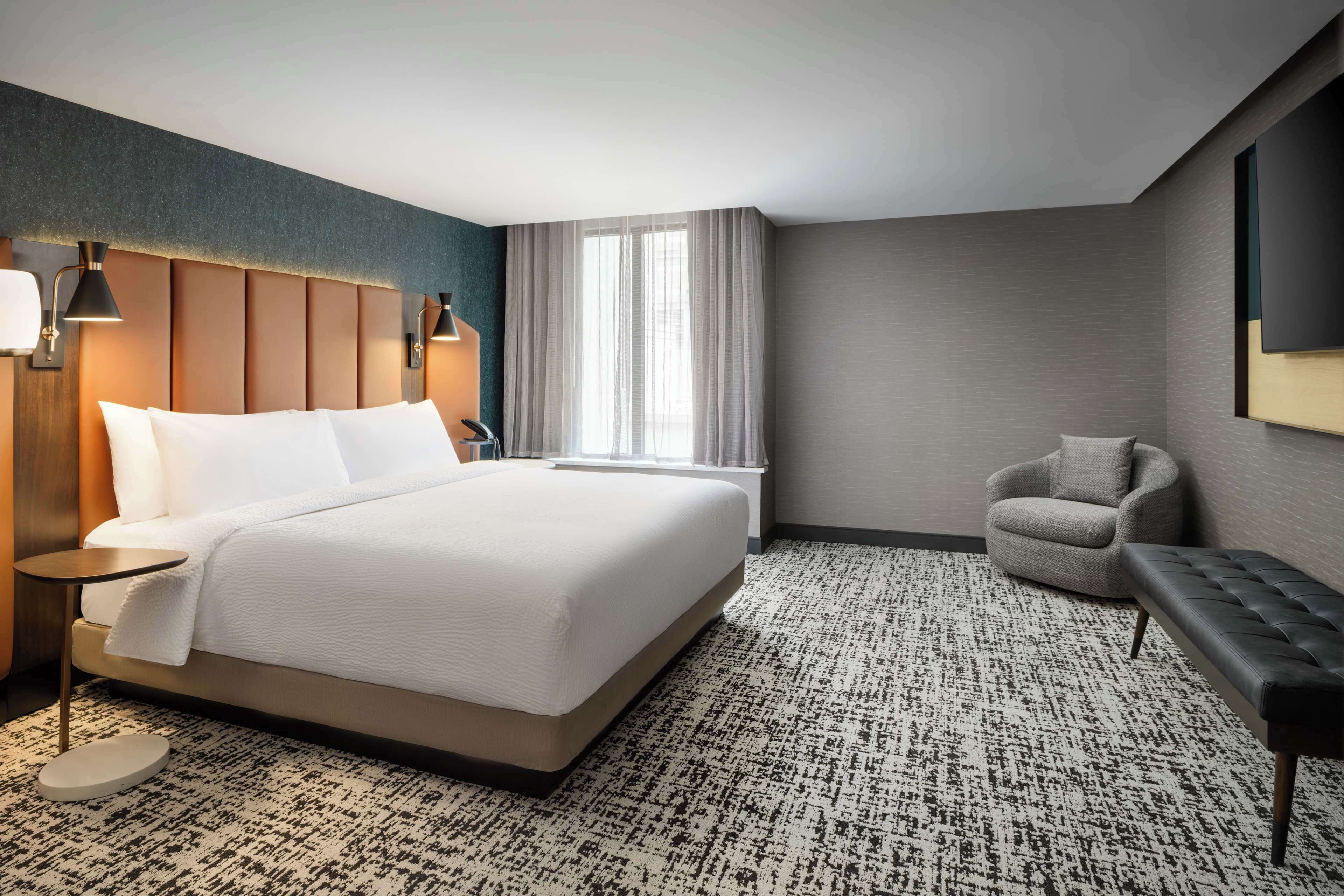 A hotel room with a large bed, a round side table, a chair, a bench, a window with curtains, and a wall-mounted TV.