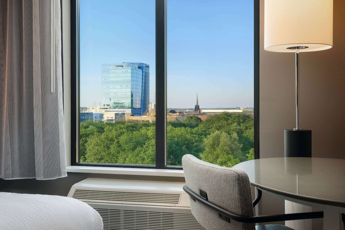 A hotel room with a large window, a round table, a chair, a lamp, and a bed.