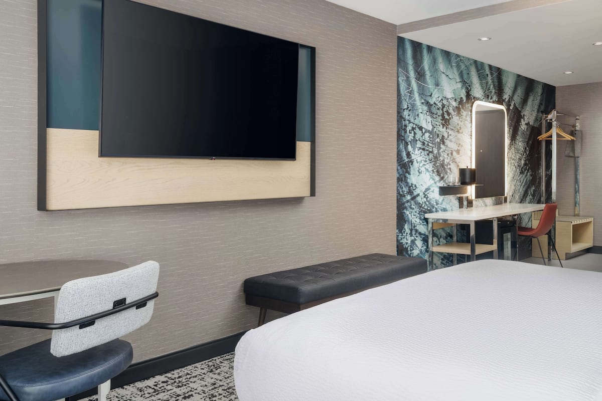 A modern hotel room with a large bed, a desk with a chair, a television mounted on the wall, and a mirror with lighting.