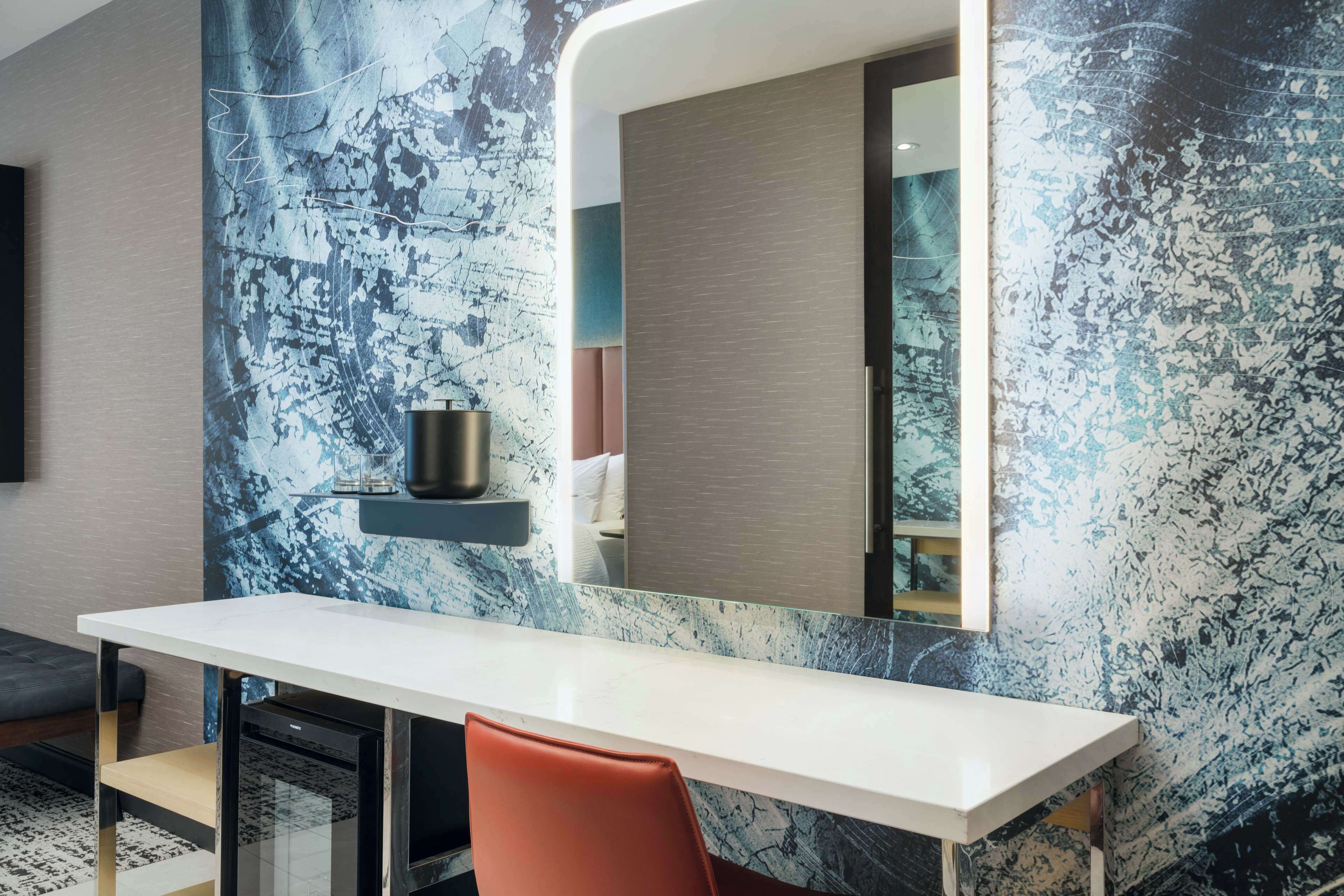 A modern hotel room with a white desk, a red chair, a large mirror, and a textured wall.