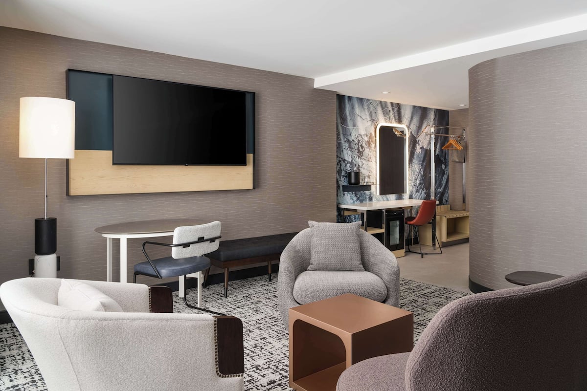 A modern hotel room with a large flat-screen TV, a dining area, a seating area with a sofa and chairs, and a large mirror.