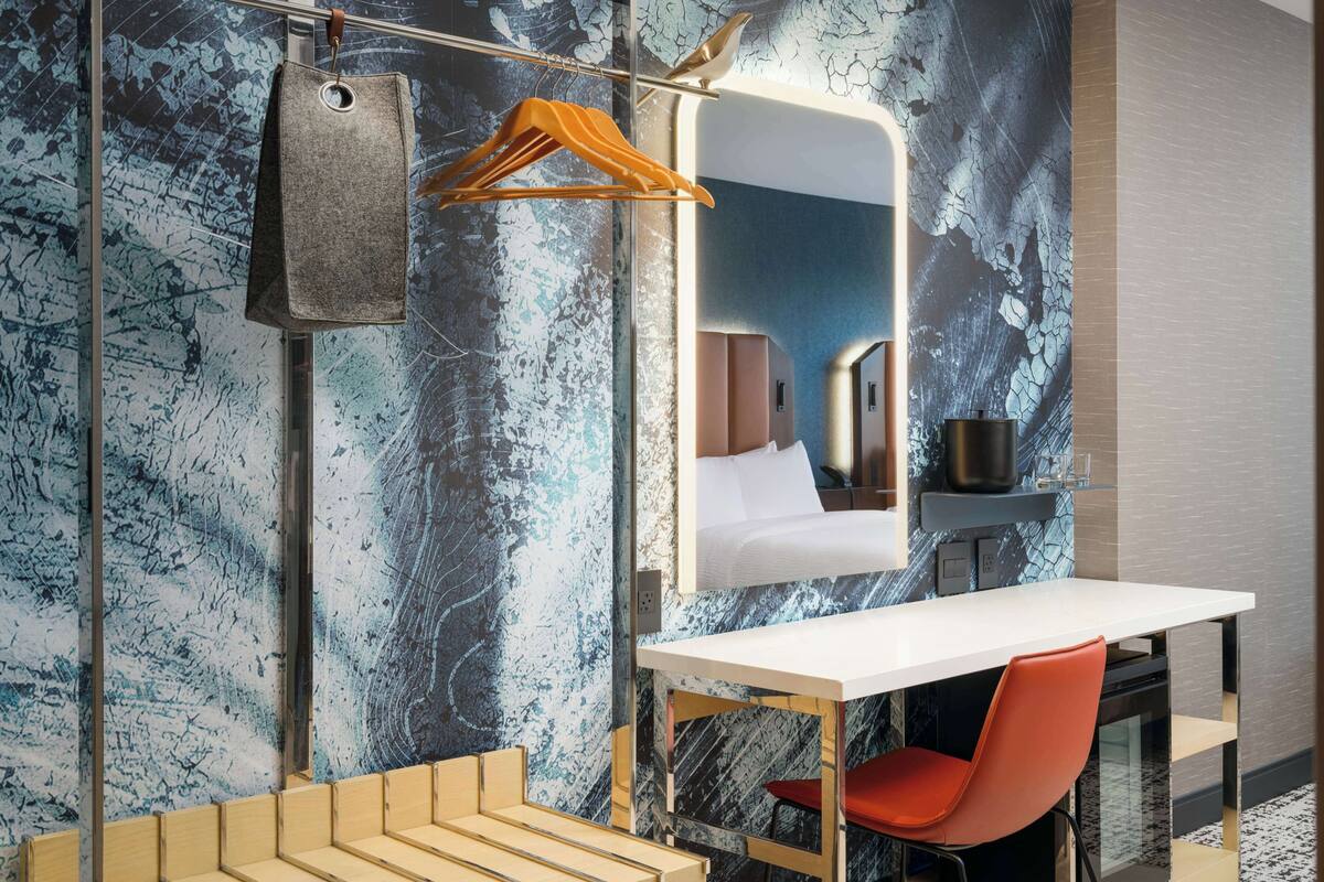 A modern hotel room with a marble-patterned wall, a white desk, a red chair, a hanging coat, and a large mirror.