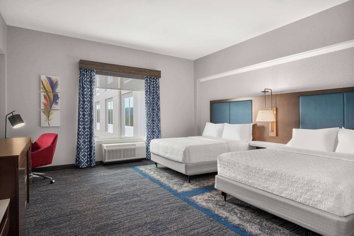 Une chambre d’hôtel avec deux lits, un bureau, une chaise rouge, une fenêtre avec des rideaux à motifs et des œuvres d’art accrochées au mur.