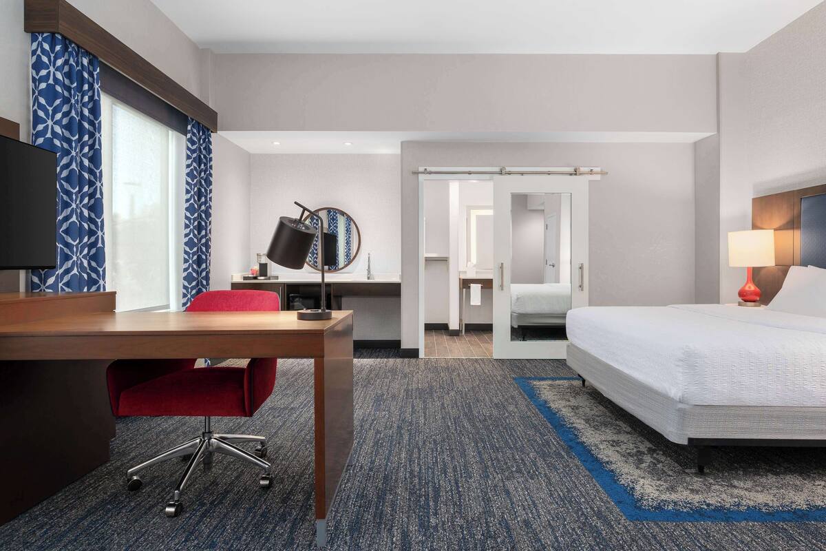 Une chambre d’hôtel avec un bureau, une chaise rouge, un lit, une télévision, un miroir et une lampe.