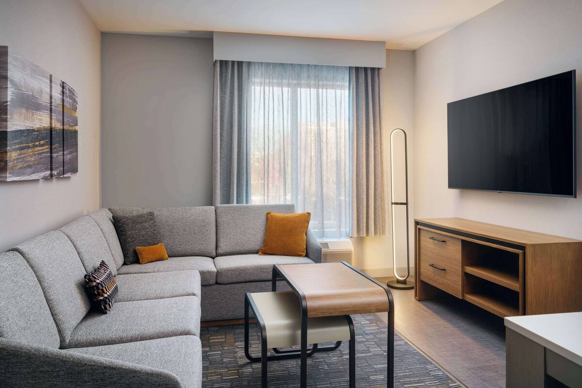 A modern hotel room with a grey sectional sofa, a wooden side table, a flat-screen TV mounted on the wall, and a large window with sheer curtains.