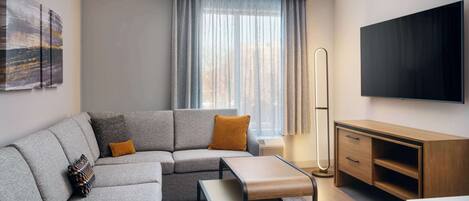 A modern hotel room with a grey sectional sofa, a wooden side table, a flat-screen TV mounted on the wall, and a large window with sheer curtains.