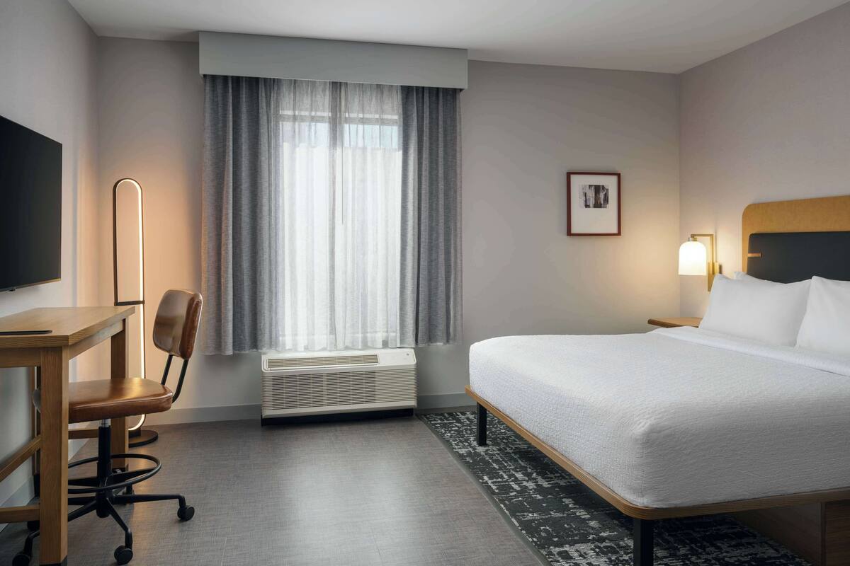 A hotel room with a bed, a desk, a chair, a TV, a window with curtains, and a framed picture on the wall.