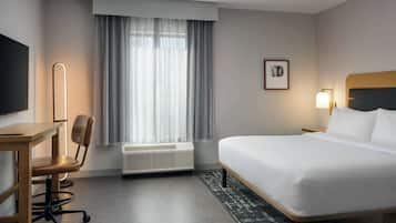 Habitación de hotel con cama, escritorio, silla, televisor, ventana con cortinas e imagen en un marco en la pared.