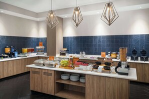 A modern breakfast bar with a variety of food items and beverages, geometric patterned backsplash, and pendant lighting. - Homewood Suites by Hilton Raleigh Downtown (Raleigh)