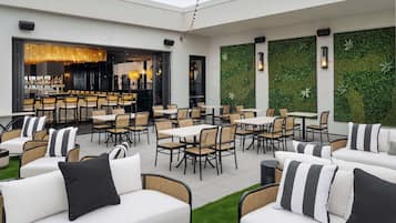 An outdoor patio area with white sofas, wicker chairs, and tables. There is a bar area with a glass enclosure and a green wall with foliage.