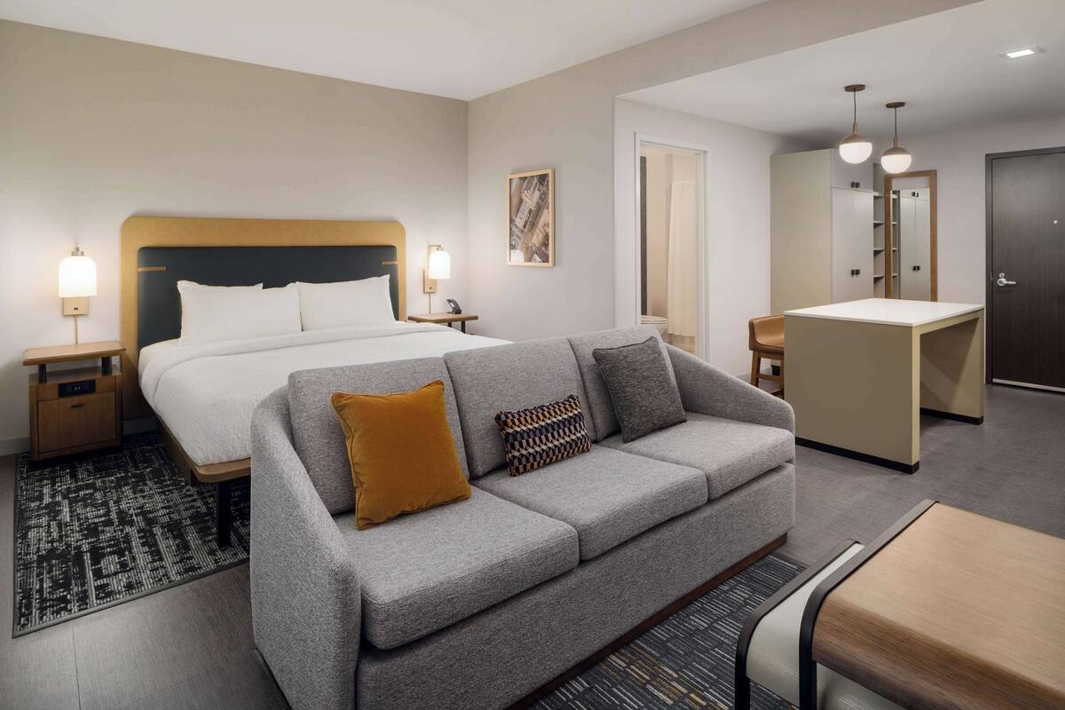 A modern hotel room with a large bed, a grey sofa, a small table, and a desk.