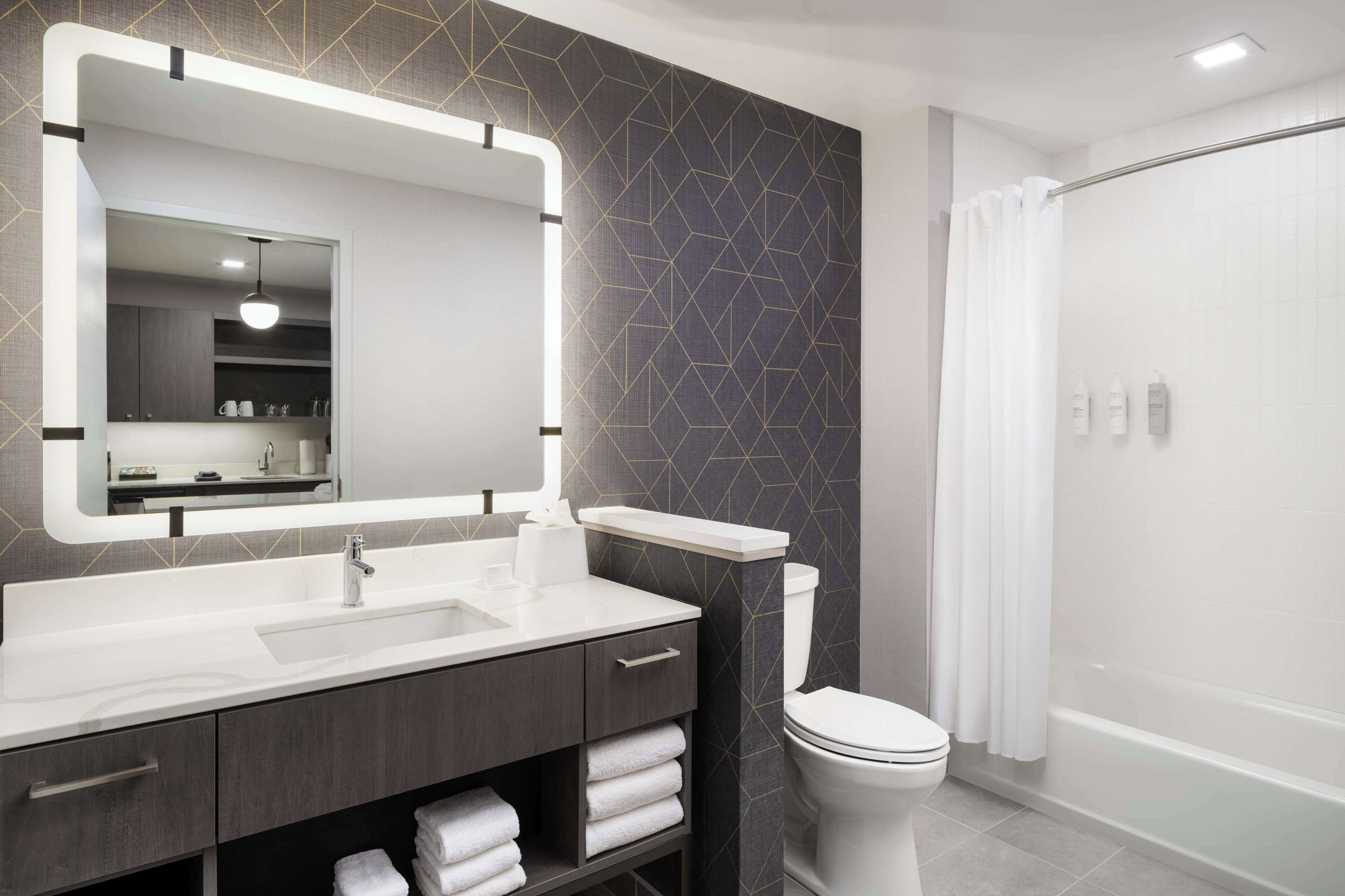 A modern bathroom with a large mirror, a white sink, dark patterned wall, and a shower area.