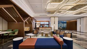 A modern hotel lobby with seating areas, a reception desk, and a view of the restaurant in the background.