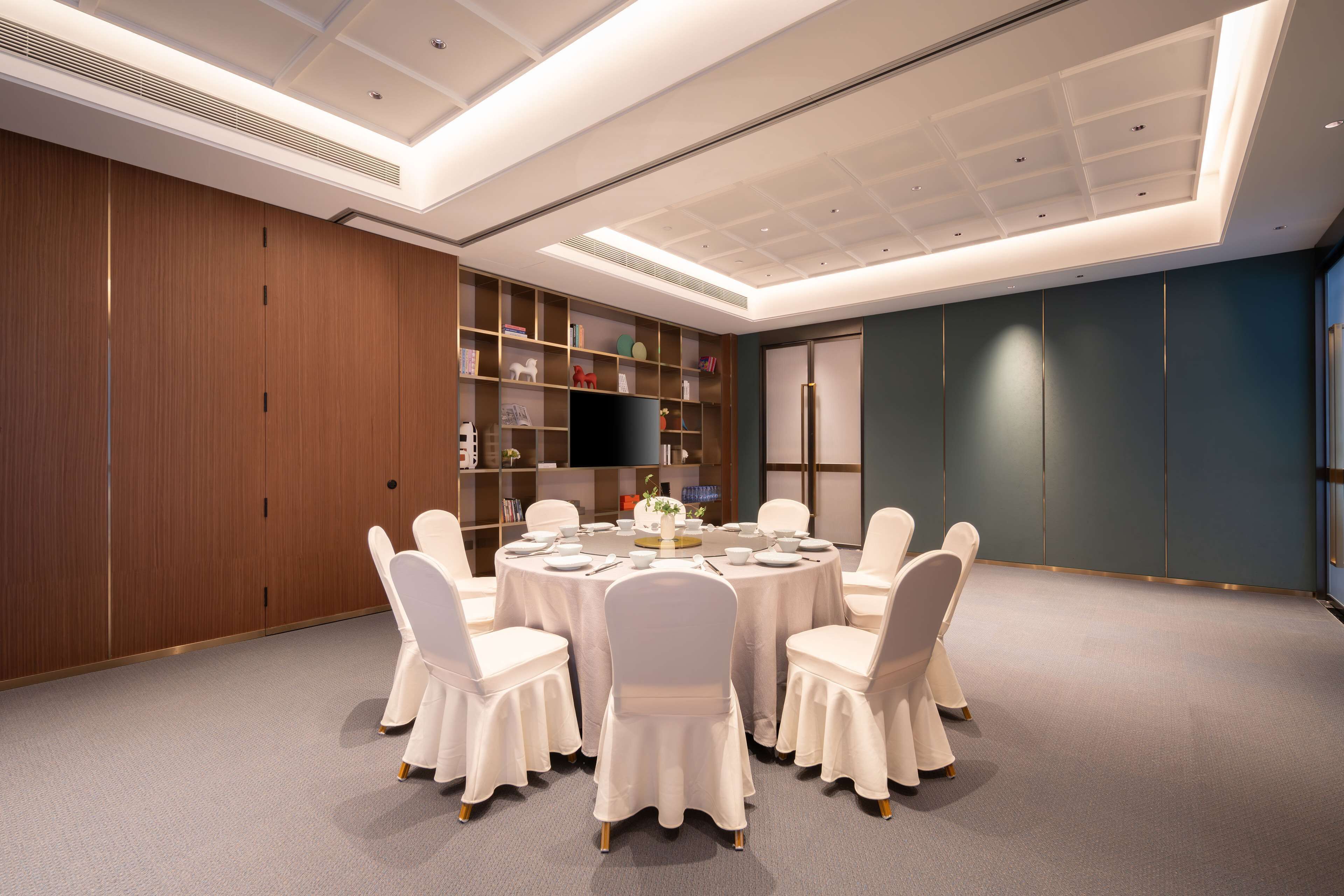 A conference room with a round table, white chairs, and a bookshelf with various items.